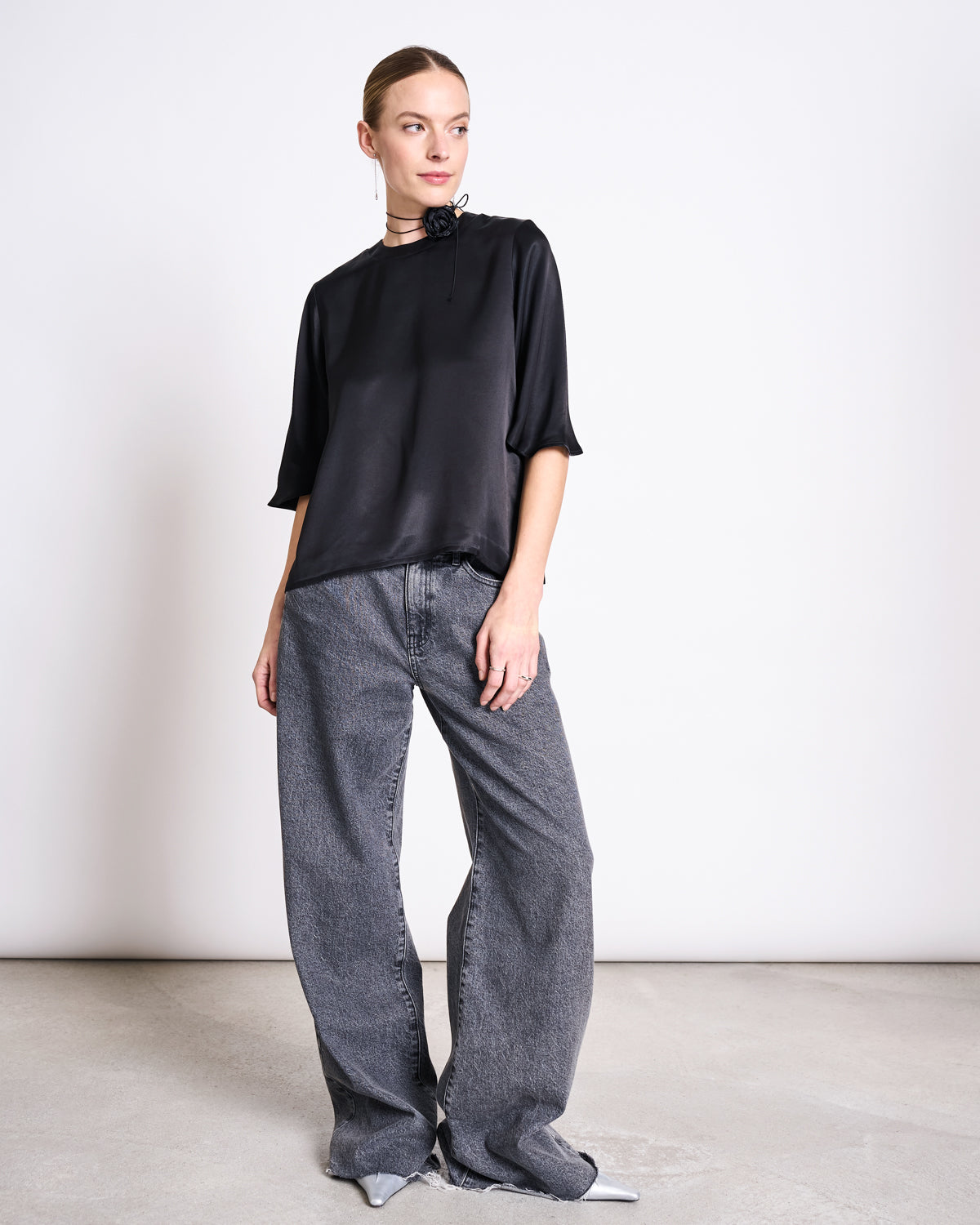 A woman stands indoors wearing the jan-n-june SILKY SHIRT NAMORA BLACK with half sleeves, paired with wide-leg gray jeans, silver pointed shoes, and a black choker. She gazes slightly left against a plain white background.