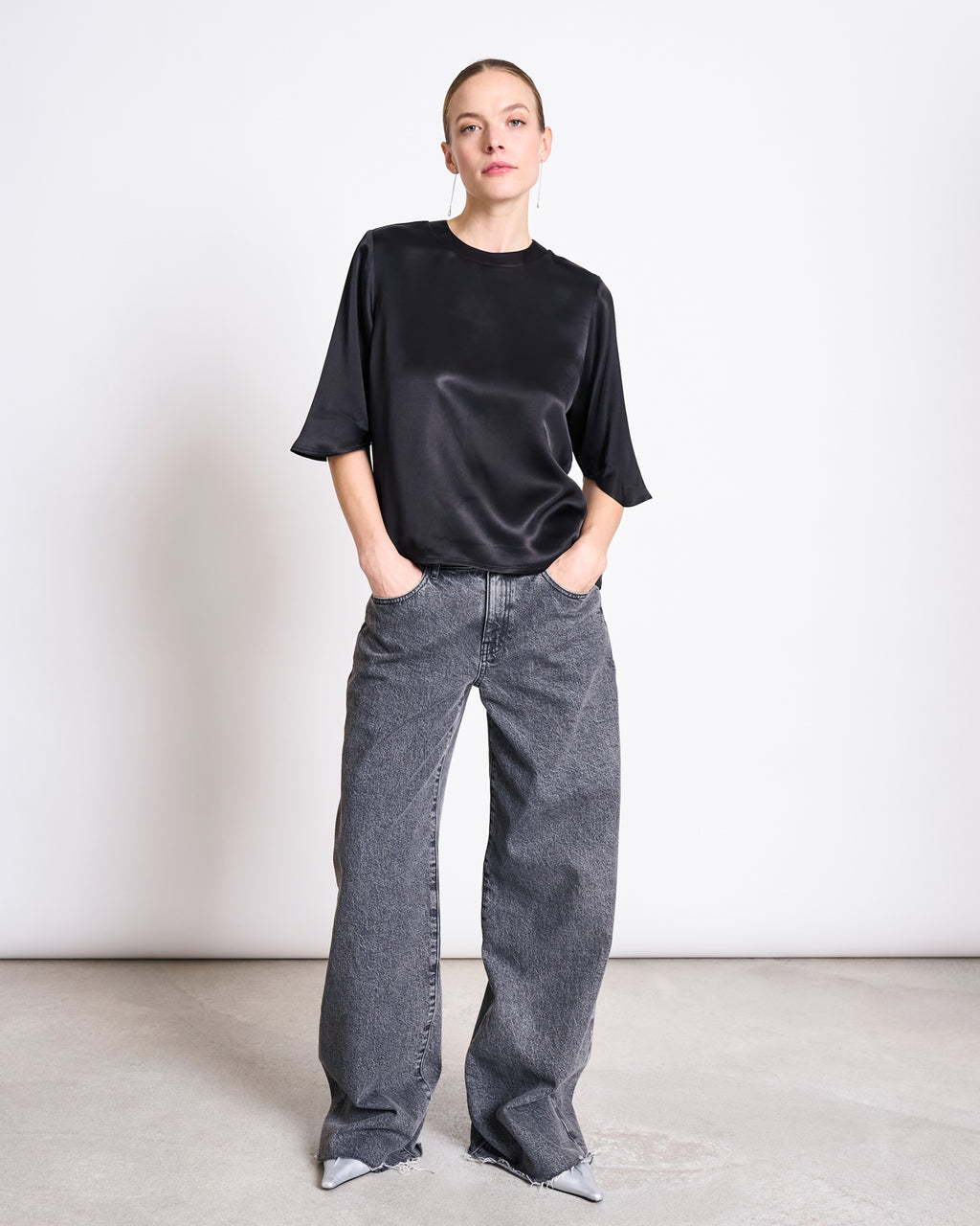 A person stands against a plain white background, wearing the jan-n-june SILKY SHIRT NAMORA BLACK with wide-leg gray jeans and pointed gray shoes, hands in pockets and a neutral expression.