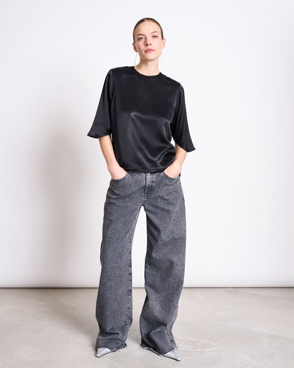A person stands against a plain white background, wearing the jan-n-june SILKY SHIRT NAMORA BLACK with wide-leg gray jeans and pointed gray shoes, hands in pockets and a neutral expression.