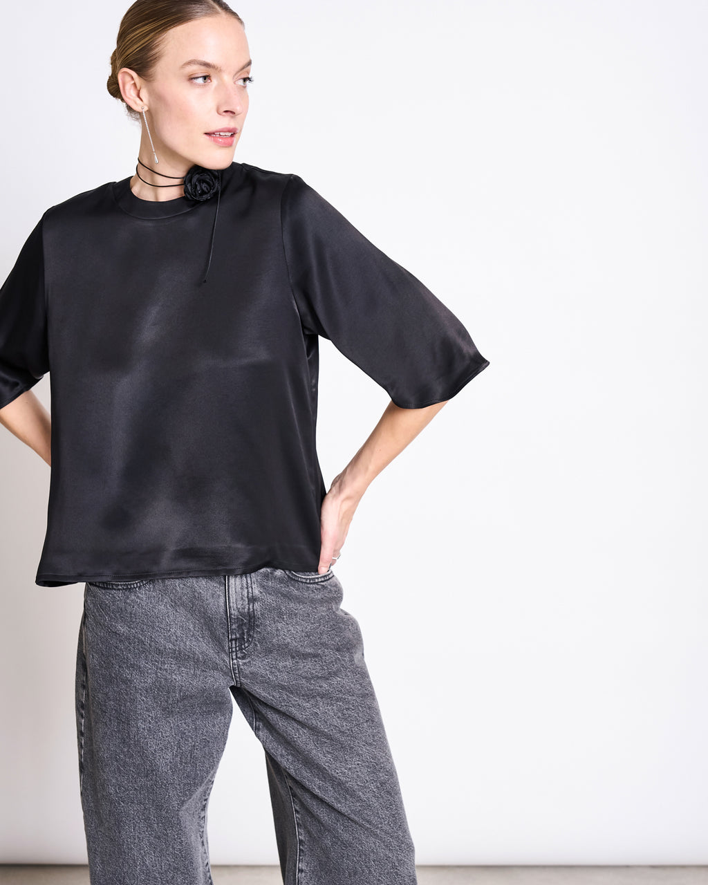 A woman stands against a white background wearing the jan-n-june SILKY SHIRT NAMORA BLACK, made with ENKA Viscose, paired with high-waisted gray jeans; she has one hand on her hip and her hair pulled back.