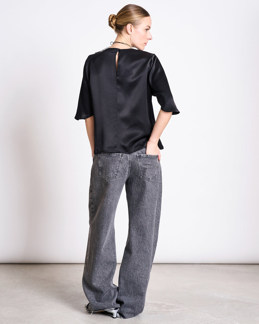 A woman faces away in a jan-n-june SILKY SHIRT NAMORA BLACK blouse with a keyhole back and wide-leg gray jeans. Her hair is in a neat bun, hands in her back pockets, set against a plain light background.