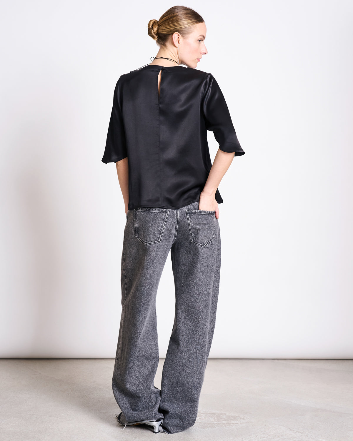 A woman faces away in a jan-n-june SILKY SHIRT NAMORA BLACK blouse with a keyhole back and wide-leg gray jeans. Her hair is in a neat bun, hands in her back pockets, set against a plain light background.