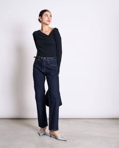 A woman stands against a plain white wall wearing the jan-n-june ASYMMETRICAL LONGSLEEVE CASMA BLACK GOTS top, paired with dark high-waisted jeans, silver pointed-toe heels, and a black belt, looking left with one arm behind her back.
