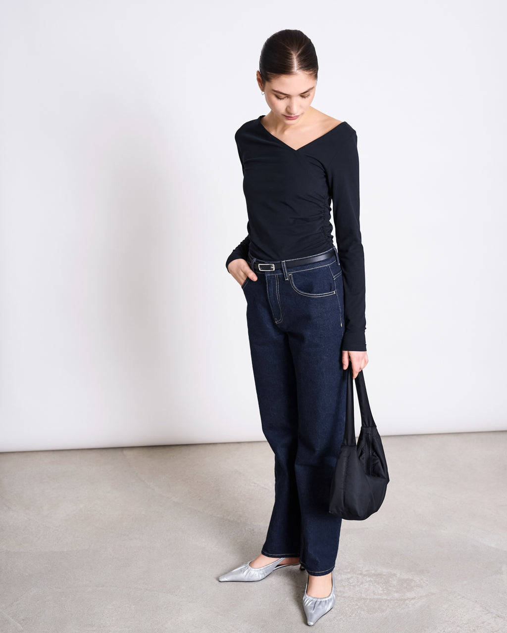 A woman stands indoors on a light floor, looking down, wearing jan-n-june’s ASYMMETRICAL LONGSLEEVE CASMA BLACK GOTS top with dark high-waisted jeans, a belt, silver pointed shoes, and holding a black handbag.