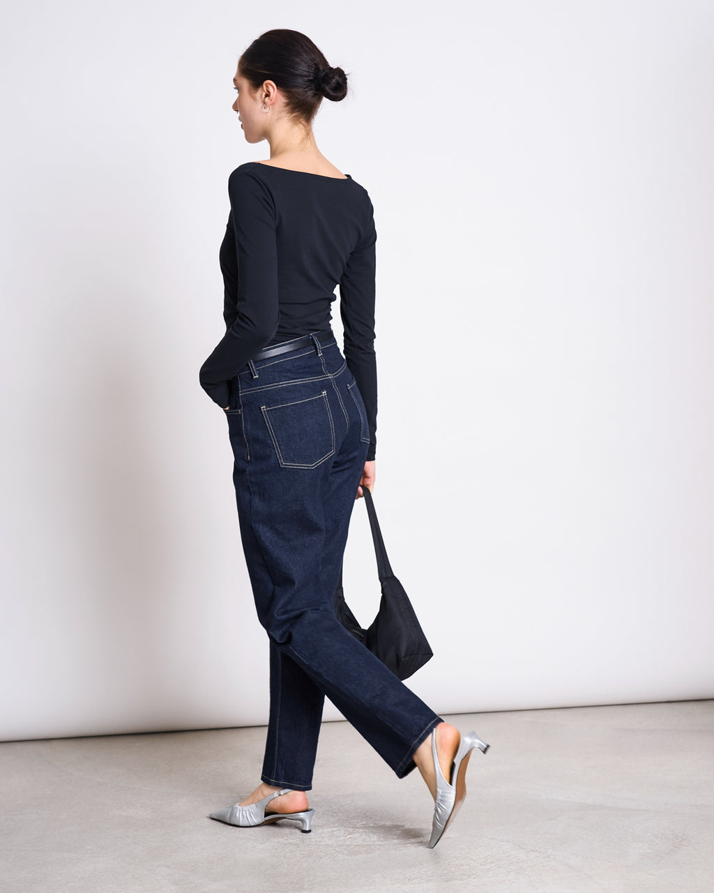 A woman with dark hair in a bun wears the jan-n-june ASYMMETRICAL LONGSLEEVE CASMA BLACK GOTS top, dark jeans, and gray heels. She carries a black purse and walks away in a minimal, light-colored room.