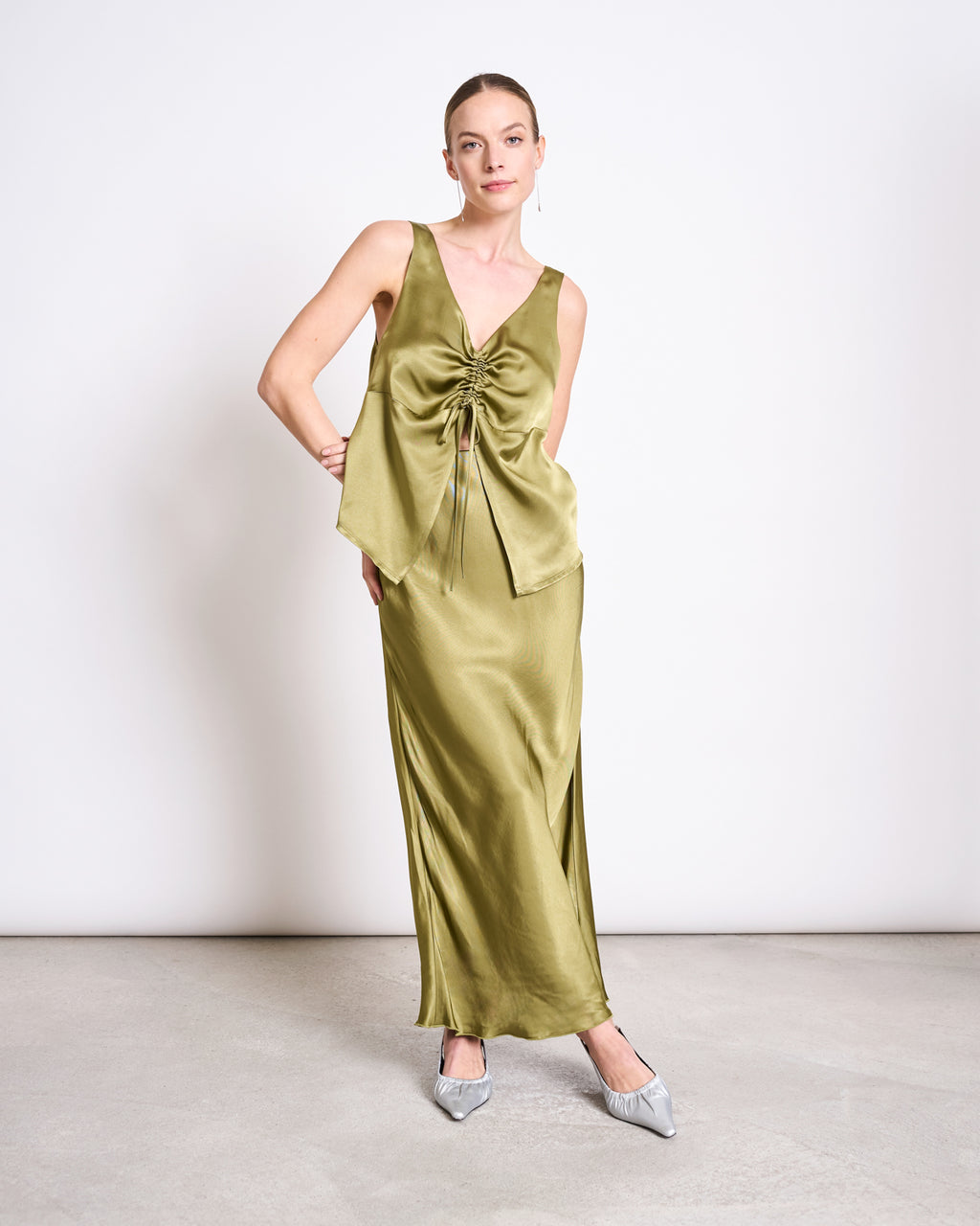 A woman stands against a white wall wearing the jan-n-june RUFFLED V-NECK TOP TALARA in woody olive, styled with her hair pulled back and gray pointed-toe shoes.