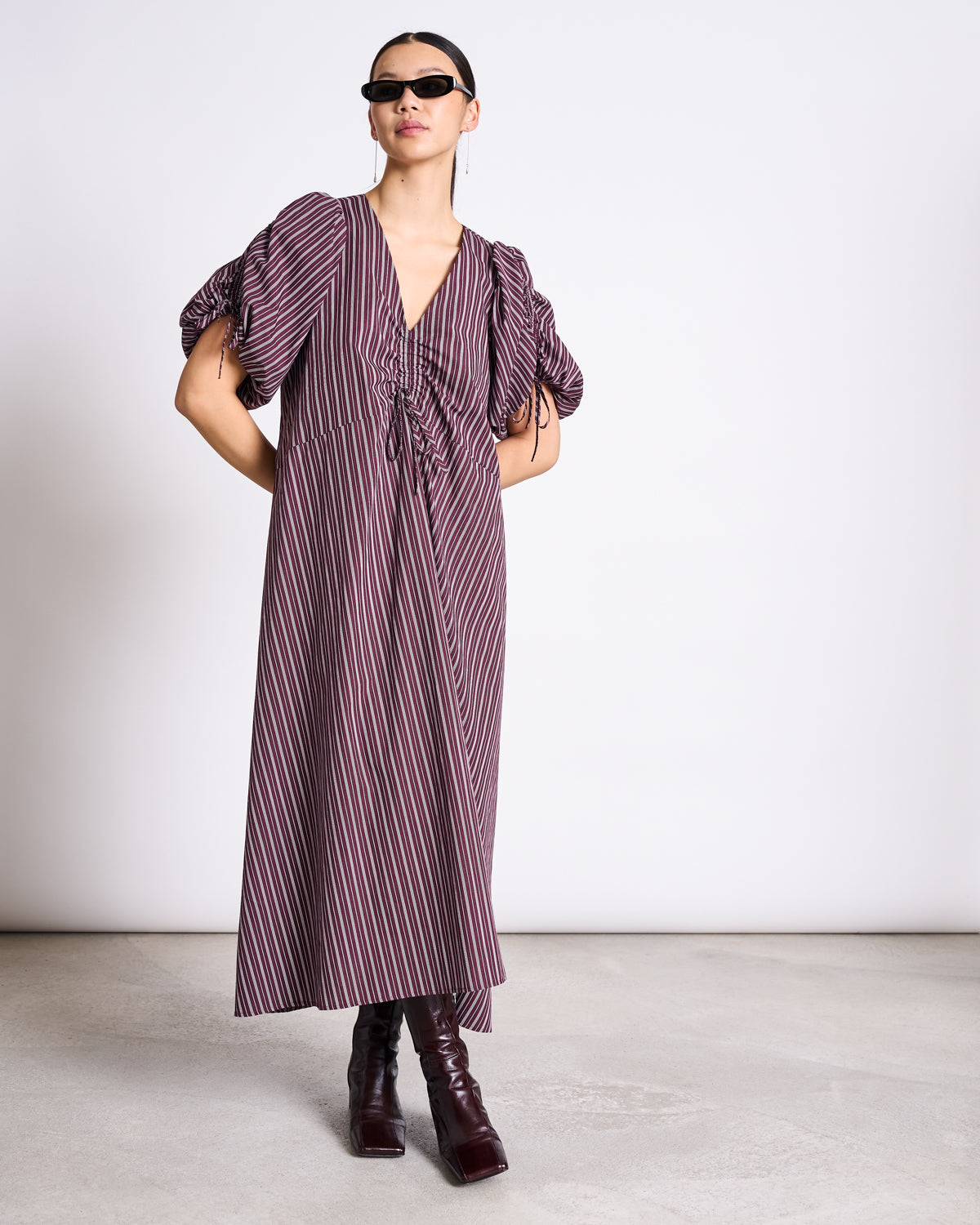 Wearing dark sunglasses and the jan-n-june MAXI DRESS DANS CHERRY LACQUER WHITE, featuring ruched sleeves and a deep V-neck, this confident individual pairs it with knee-high heeled boots for an elegant look.