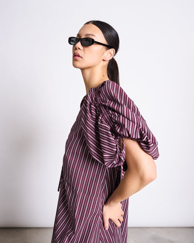 A woman in dark sunglasses wears the jan-n-june MAXI DRESS DANS CHERRY LACQUER WHITE, featuring maroon and white vertical stripes, as she stands in profile against a plain light background with her hair pulled back.