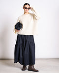 A person wearing black sunglasses, a jan-n-june OVERSIZED ALPACA MIX JUMPER WOOLY SONA OFFWHITE, a long black skirt, brown boots, and holding a black clutch bag stands against a plain background.