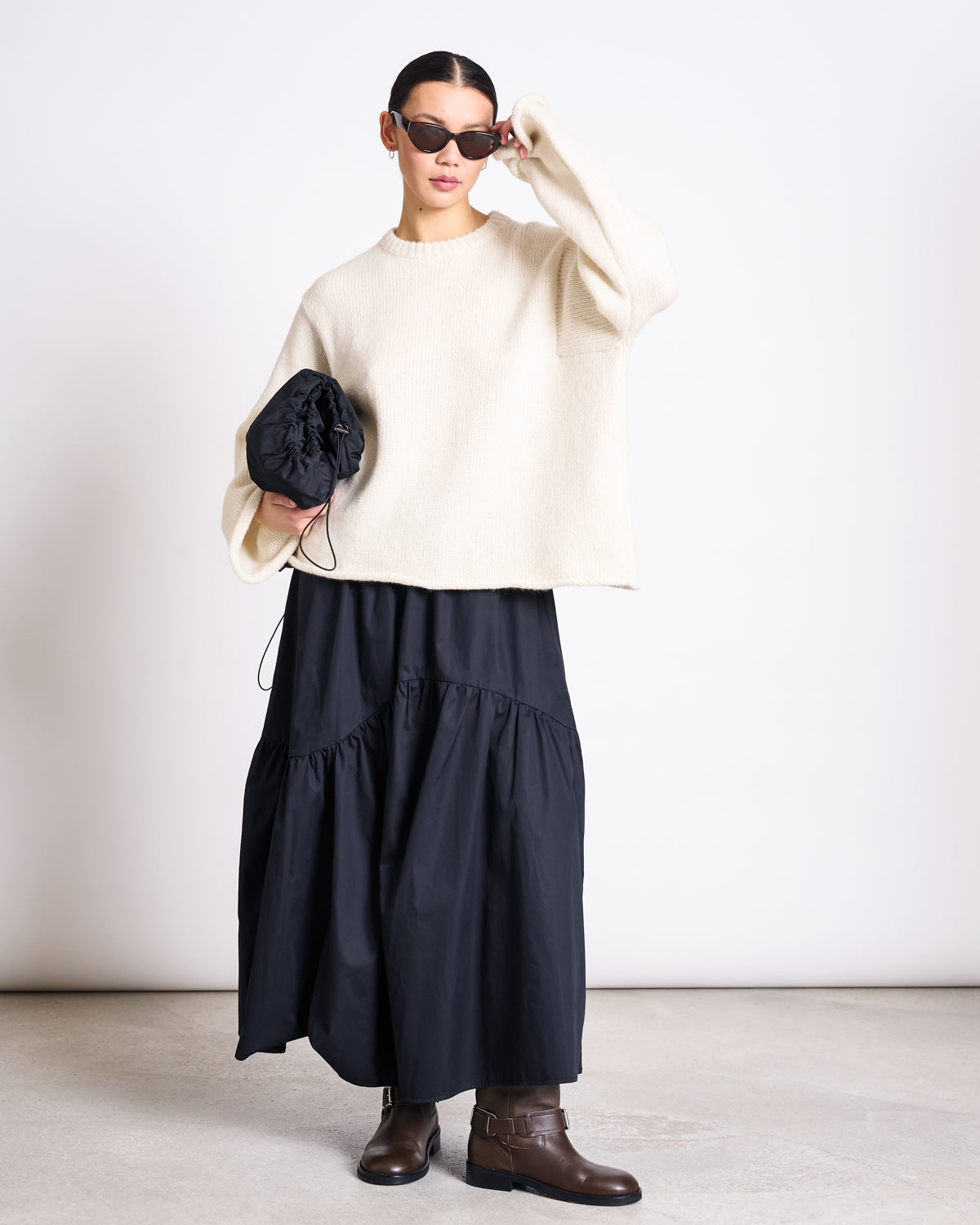 A person wearing black sunglasses, a jan-n-june OVERSIZED ALPACA MIX JUMPER WOOLY SONA OFFWHITE, a long black skirt, brown boots, and holding a black clutch bag stands against a plain background.