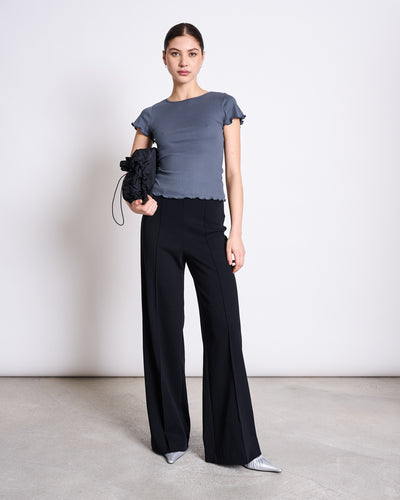 A woman stands against a plain background wearing jan-n-junes WIDE PANTS LAREDO BLACK GOTS, paired with a short-sleeved gray top, gray pointed shoes, and holding a black bag with a floral detail.
