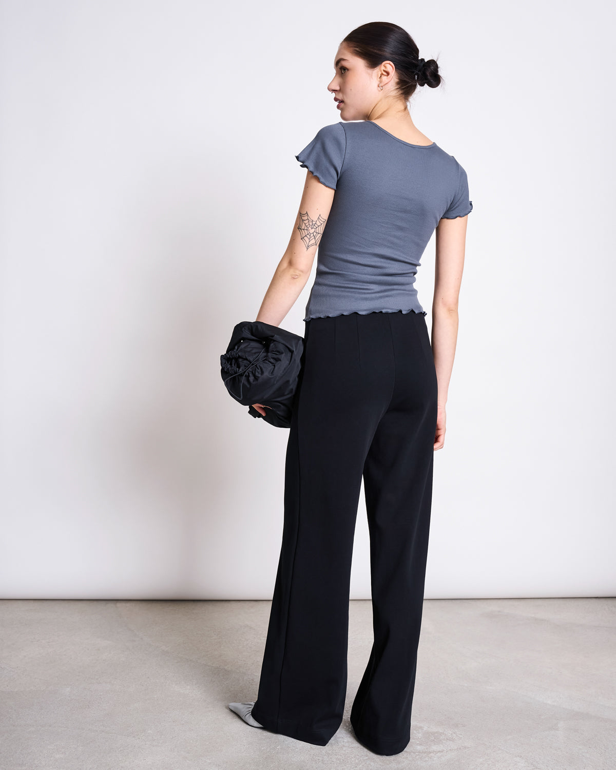 A woman with her hair in a bun, showing a tattoo on her upper left arm, wears the jan-n-june MINIRIB T-SHIRT REN TURBULENCE GOTS in gray and black wide-leg pants. She holds a black bag against a plain light background.