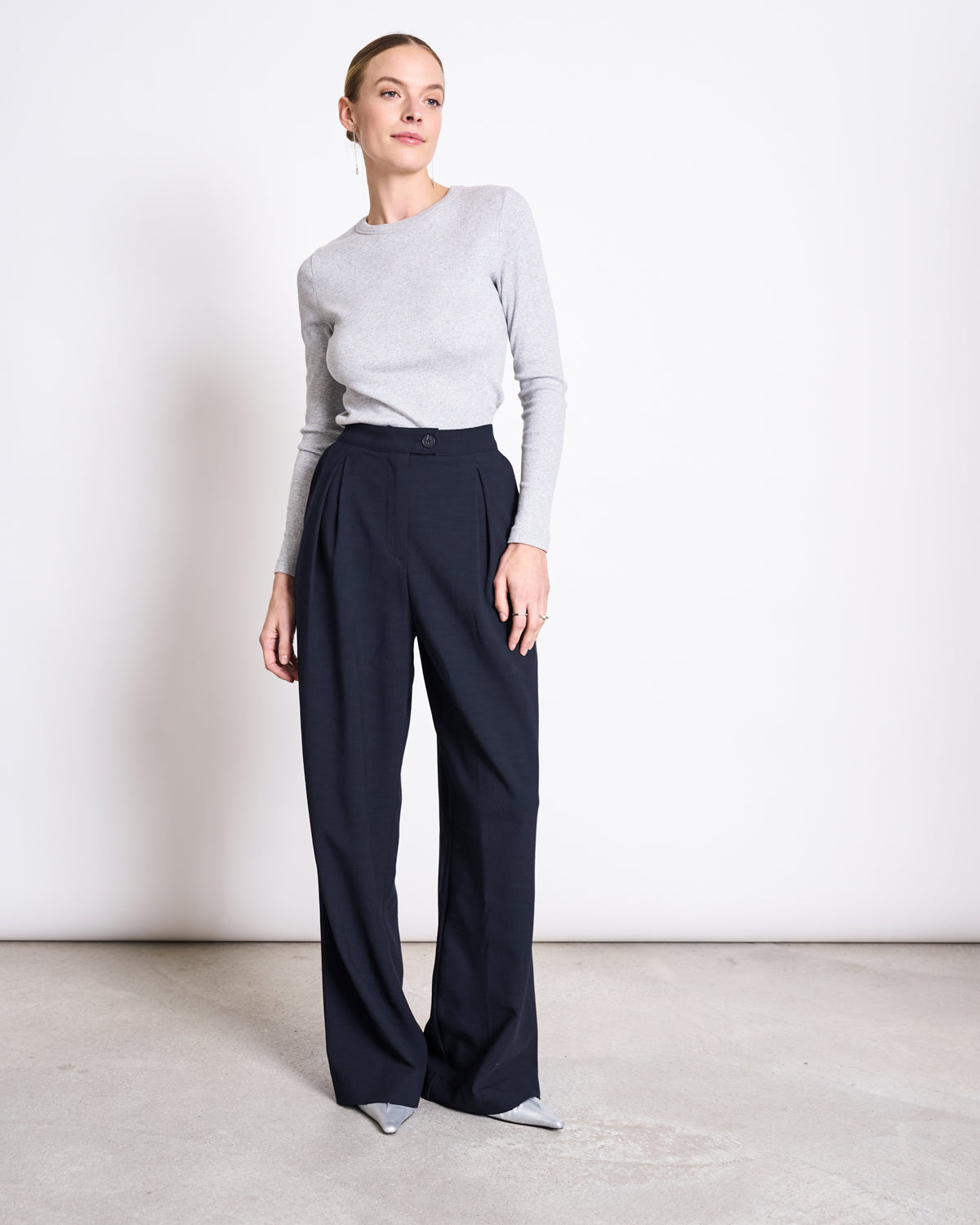 A woman stands against a plain white wall, wearing a fitted light gray long-sleeve top tucked into high-waisted jan-n-june PLEATED WIDE PANTS NIVA NIGHTFALL and gray pointed-toe shoes. Her hair is pulled back, and she poses with one hand by her side.
