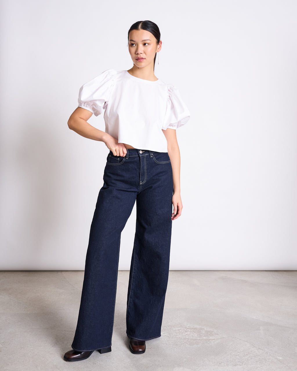 A woman stands in a minimal studio wearing jan-n-june’s WIDE LEG LOWRISE JEANS TACNA DARK BLUE DENIM GOTS and a white puff-sleeve blouse with black heels, posing with one hand on her hip and gazing slightly to the side.