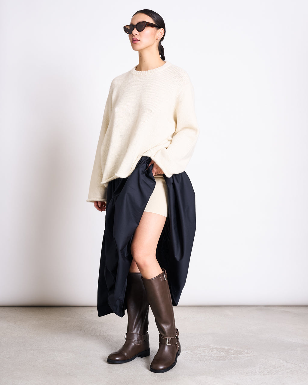 A woman in dark sunglasses, cream shorts, knee-high brown boots, and a black coat draped over her arm wears the jan-n-june OVERSIZED ALPACA MIX JUMPER WOOLY SONA OFFWHITE, standing against a plain white wall.