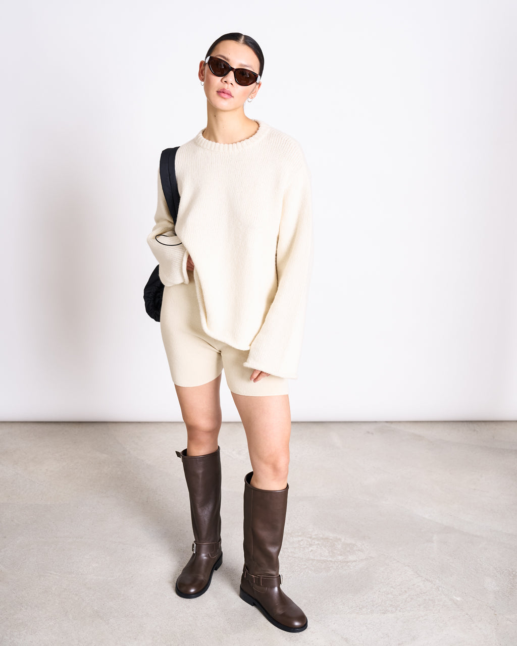 A woman in sunglasses and the jan-n-june OVERSIZED ALPACA MIX JUMPER WOOLY SONA OFFWHITE, paired with cream shorts, dark knee-high boots, and a black shoulder bag, stands against a plain light background.
