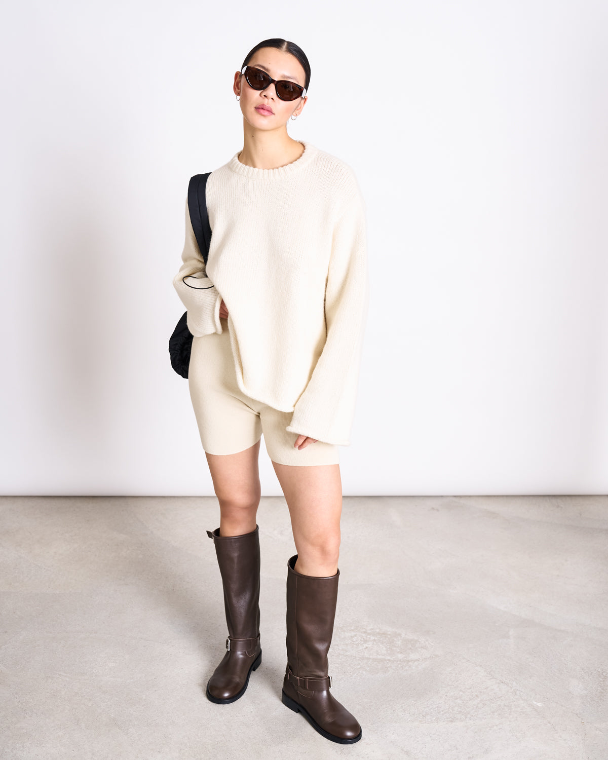 A woman in sunglasses and the jan-n-june OVERSIZED ALPACA MIX JUMPER WOOLY SONA OFFWHITE, paired with cream shorts, dark knee-high boots, and a black shoulder bag, stands against a plain light background.