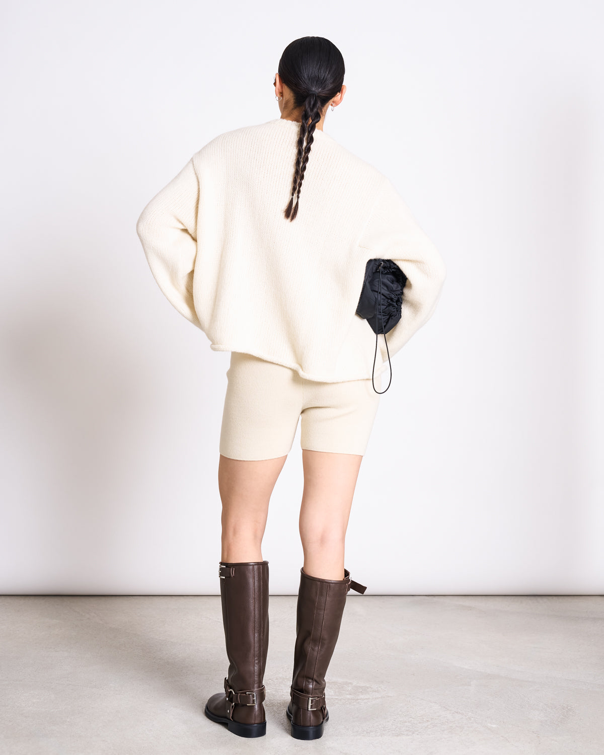 Facing away against a white background, a person with a long braid wears jan-n-junes OVERSIZED ALPACA MIX JUMPER WOOLY SONA OFFWHITE, matching cream shorts, knee-high brown boots, and holds a small black bag.