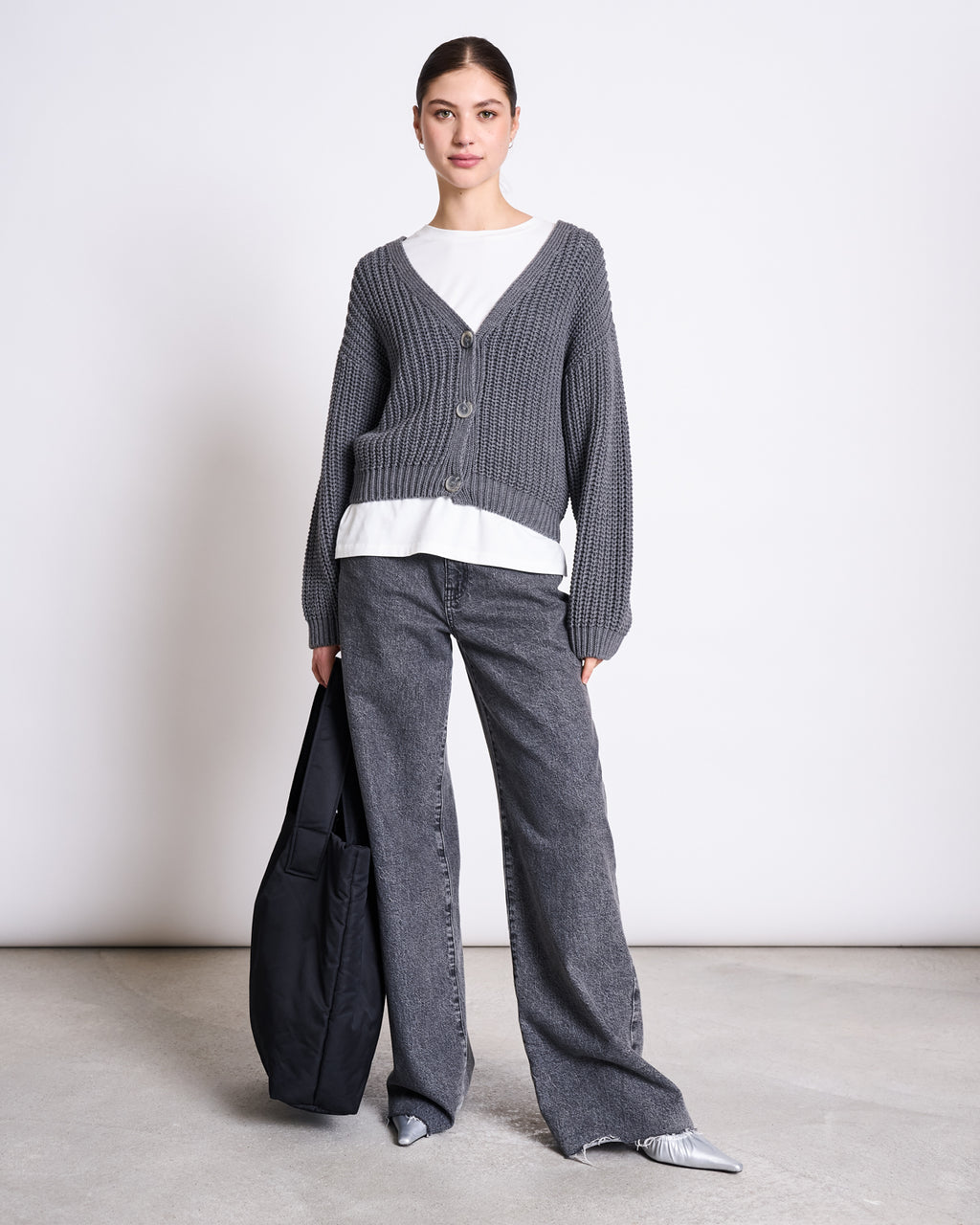 A woman stands against a plain background wearing the jan-n-june CHUNKY CARDIGAN LENA DARK GREY GOTS over a white top, paired with loose gray jeans, silver pointed shoes, and holding a black tote bag.