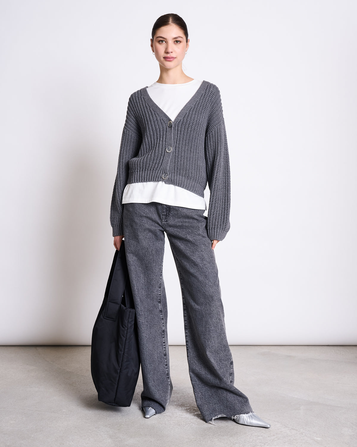A woman stands against a plain background wearing the jan-n-june CHUNKY CARDIGAN LENA DARK GREY GOTS over a white top, paired with loose gray jeans, silver pointed shoes, and holding a black tote bag.