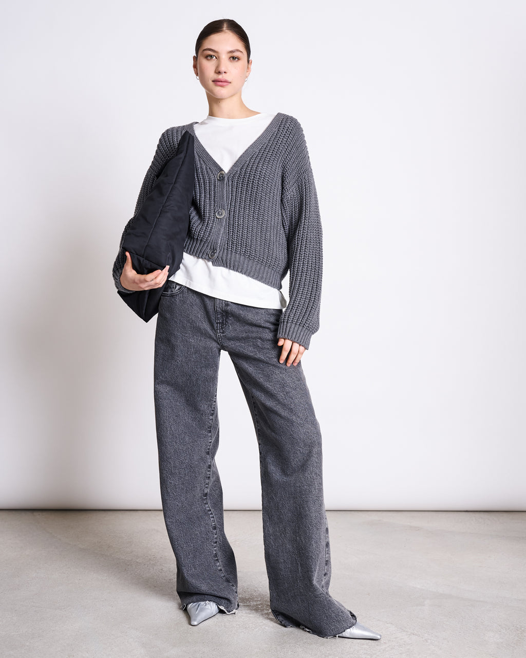 A woman stands against a plain background wearing the jan-n-june CHUNKY CARDIGAN LENA DARK GREY GOTS over a white shirt, paired with loose dark gray jeans, silver pointed shoes, and holding a black bag under her arm.