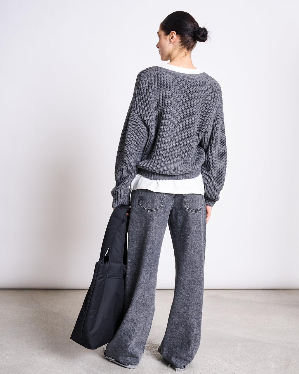 A person with dark hair in a bun, wearing the jan-n-june CHUNKY CARDIGAN LENA DARK GREY GOTS, a white shirt, and wide-leg gray jeans, stands facing away while holding a large black tote bag by their side.