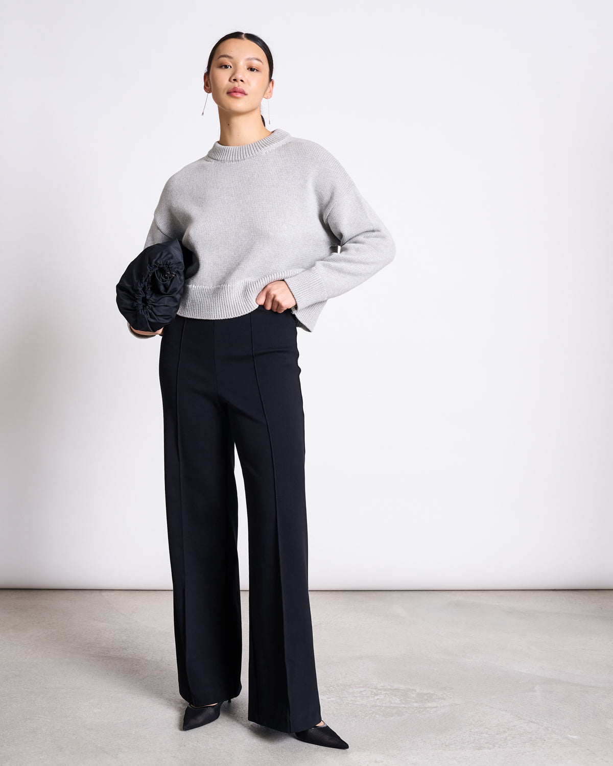 A woman stands against a plain background wearing a light gray sweater and jan-n-junes WIDE PANTS LAREDO BLACK GOTS—high-waisted wide-leg black pants made from organic cotton—paired with pointed black shoes and holding a black clutch.