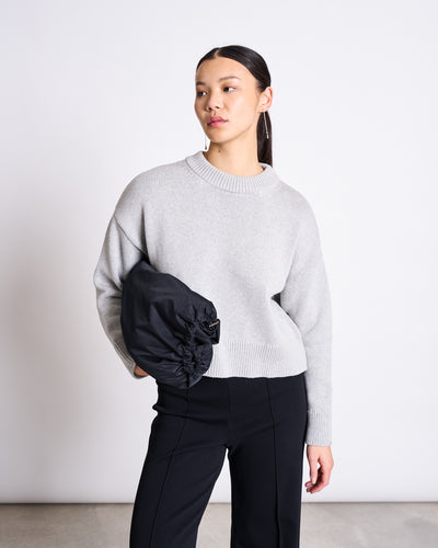 A woman with straight dark hair tied back wears the jan-n-june JUMPER YAGMUR LIGHT GREY GOTS in a knitted design and black pants, holding a black jacket under her left arm while standing against a plain white background.