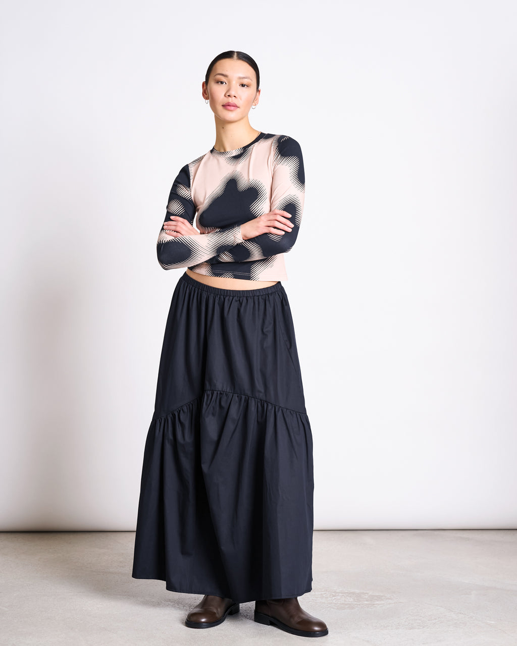 A woman stands against a white wall, arms crossed, wearing the jan-n-june LONGSLEEVE BARID DIGITAL FLOWER PRINT GOTS top and a long black tiered skirt, paired with brown boots.