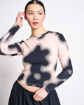 A woman with dark hair in a sleek bun wears the jan-n-june LONGSLEEVE BARID DIGITAL FLOWER PRINT GOTS top, featuring abstract black and beige patterns, paired with a dark skirt. She gazes to the side, hand near her head, against a plain background.