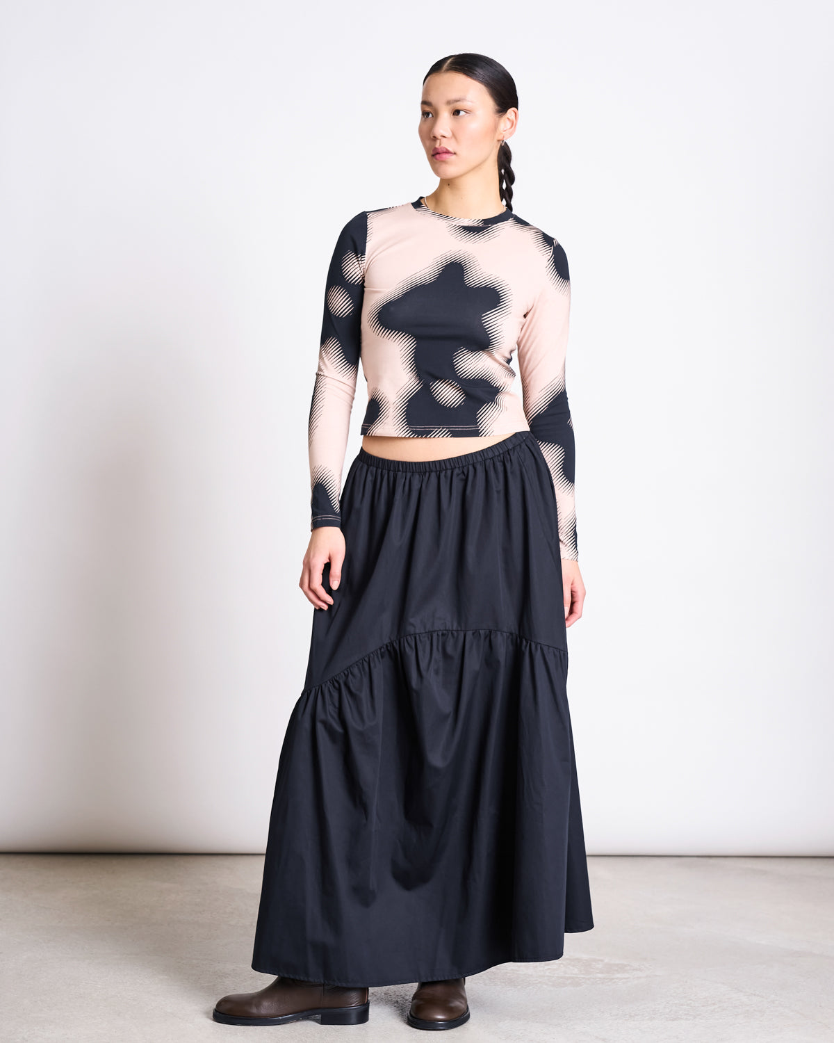 A woman stands in a minimalist room wearing the jan-n-june LONGSLEEVE BARID DIGITAL FLOWER PRINT GOTS, styled with a long black tiered skirt and dark flats. Her hair is braided as she gazes off to the side.