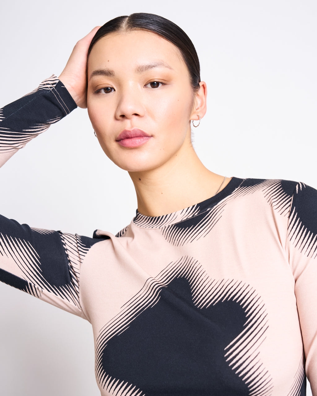 A woman with dark hair pulled back poses confidently in the jan-n-june LONGSLEEVE BARID DIGITAL FLOWER PRINT GOTS, a beige and black organic cotton jersey top, with one hand on her head against a plain background.