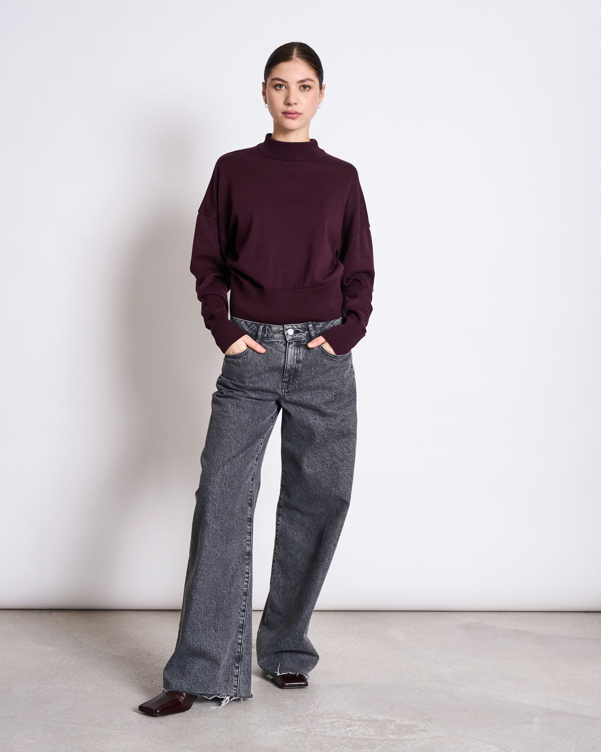 A woman stands against a white wall, wearing the jan-n-june JUMPER YIN CHERRY LACQUER GOTS with wide-leg gray jeans and dark shoes. Her hands are in her pockets as she looks confidently at the camera.