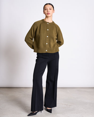 A woman stands on a concrete floor against a white wall, wearing the jan-n-june RELAXED CREWNECK CARDIGAN SIVIA WOODY OLIVE GOTS, black high-waisted wide-leg pants, and black pointed-toe heels with her hands in her pockets.