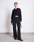 A woman with dark hair pulled back stands against a light background, wearing jan-n-june’s CRINKLE SHIRT ISTED BLACK and matching wide-leg black pants, paired with black shoes.