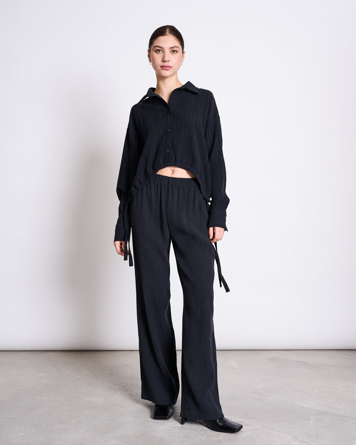 A woman with dark hair pulled back stands against a light background, wearing jan-n-june’s CRINKLE SHIRT ISTED BLACK and matching wide-leg black pants, paired with black shoes.