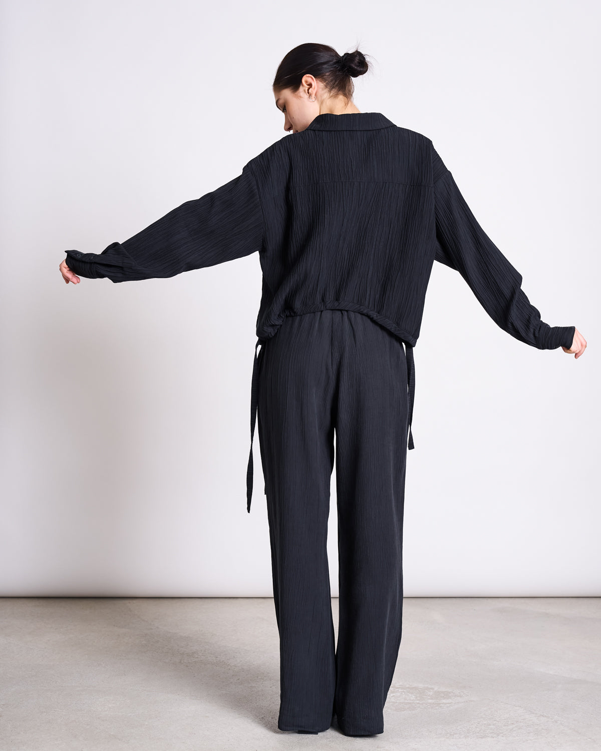 A person with dark hair in a bun stands indoors with their back to the camera, wearing jan-n-junes CRINKLE SHIRT ISTED BLACK, a loose-fitting long-sleeve top with dropped shoulders, paired with black pants against a plain background.