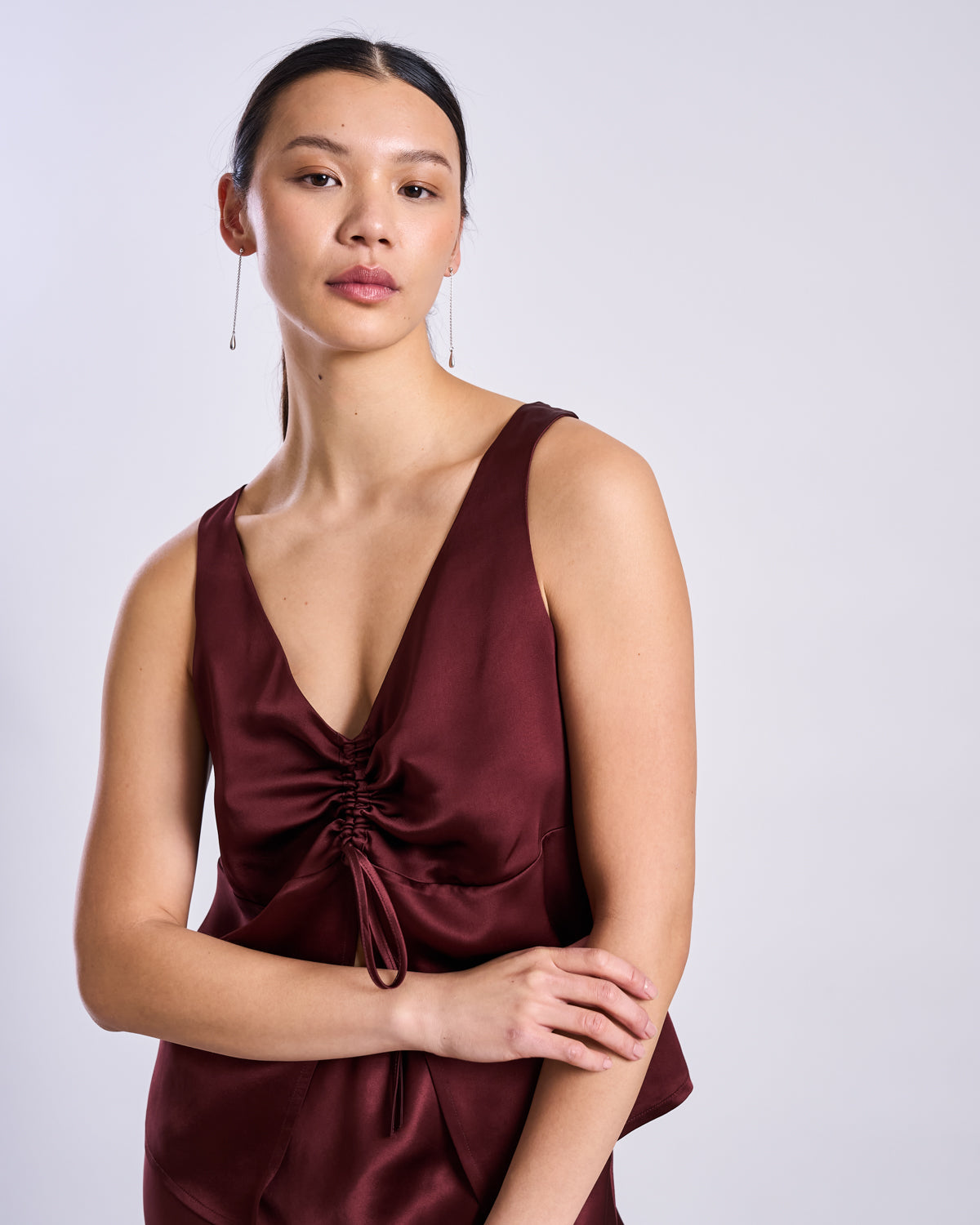 A person with long dark hair wears the jan-n-june RUFFLED V-NECK TOP TALARA in intense rust, featuring a ruched front tie, standing against a plain light background and gazing calmly at the camera for an elegant touch.