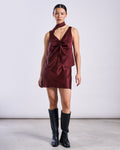 A woman stands against a plain light backdrop wearing a sleeveless jan-n-june PIURA dress in intense rust with a front tie detail, paired with the matching SILKY SCARF PIURA INTENSE RUST and black knee-high boots.