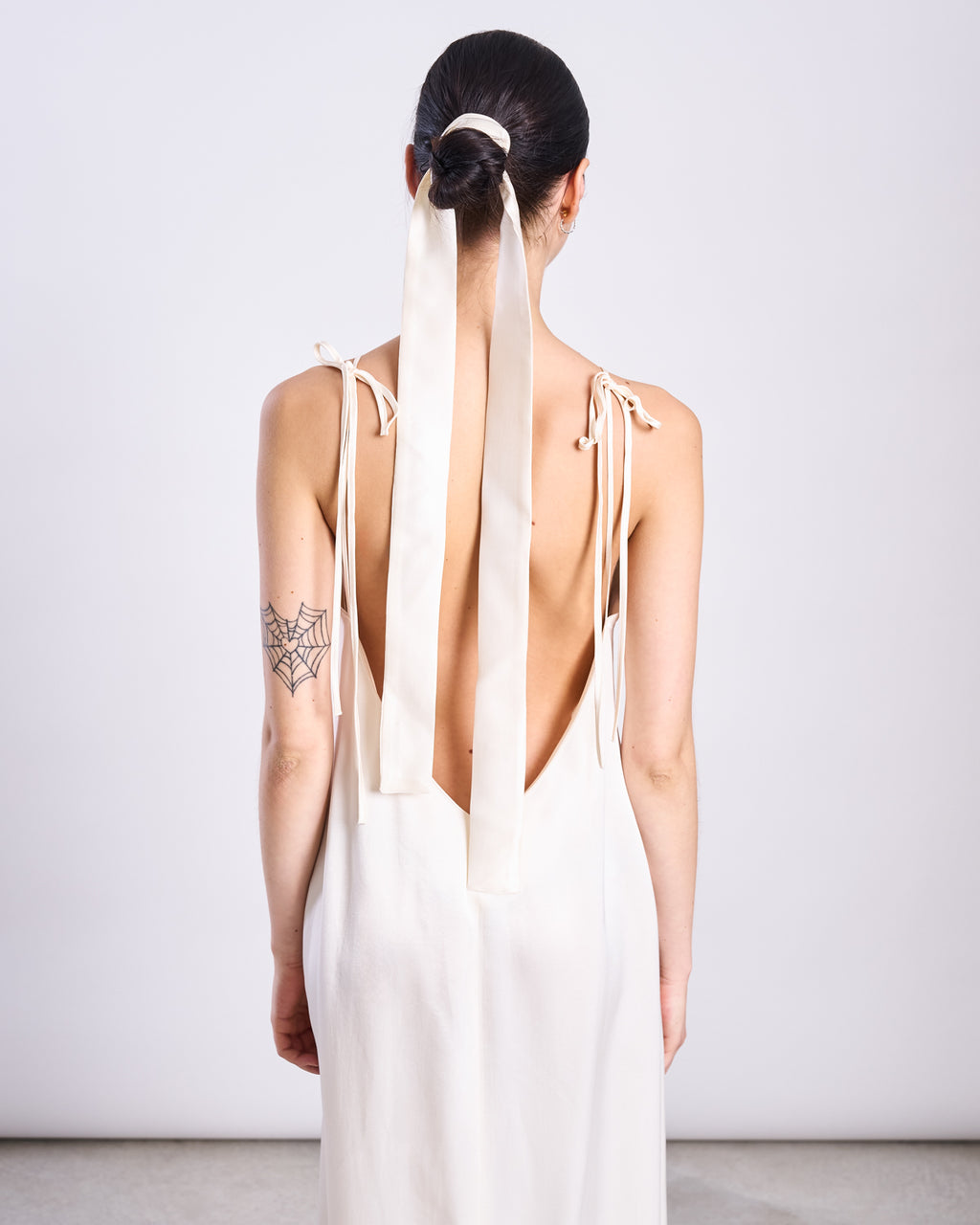 A woman with dark hair in a ponytail tied with a long, elegant jan-n-june SILKY SCARF PIURA OFFWHITE wears a sleeveless, backless ivory dress. She has a black spider web tattoo on her upper left arm and faces away from the camera.
