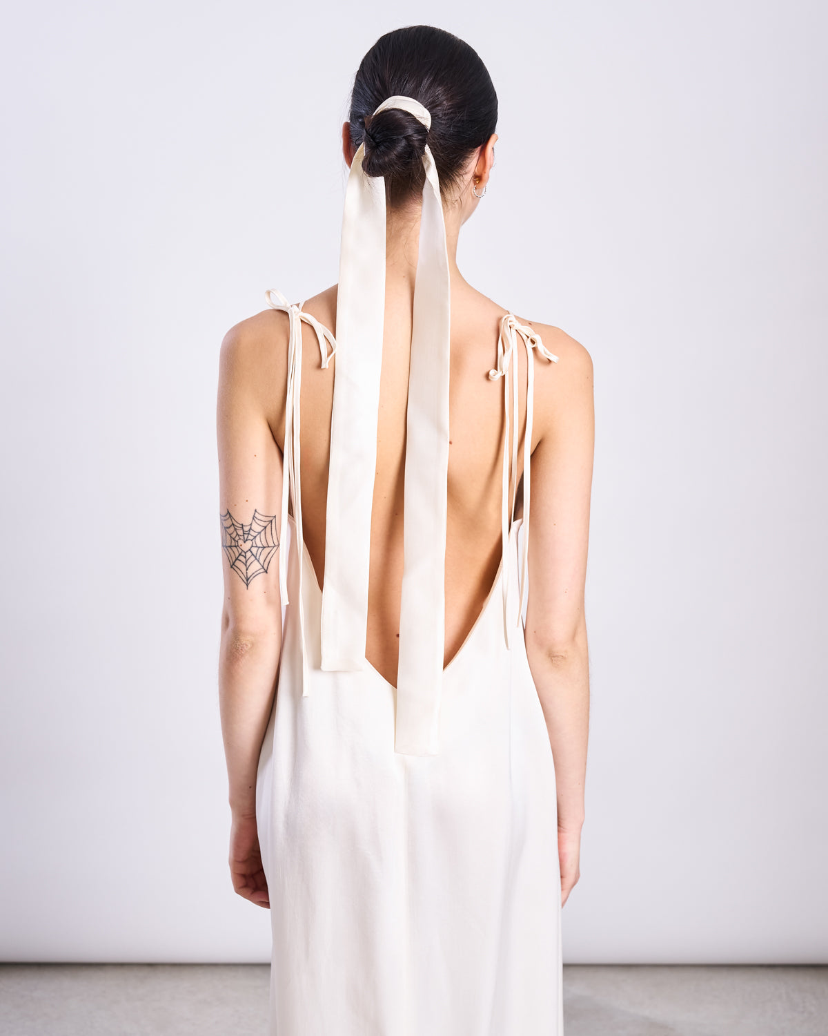 A woman with dark hair in a ponytail tied with a long, elegant jan-n-june SILKY SCARF PIURA OFFWHITE wears a sleeveless, backless ivory dress. She has a black spider web tattoo on her upper left arm and faces away from the camera.