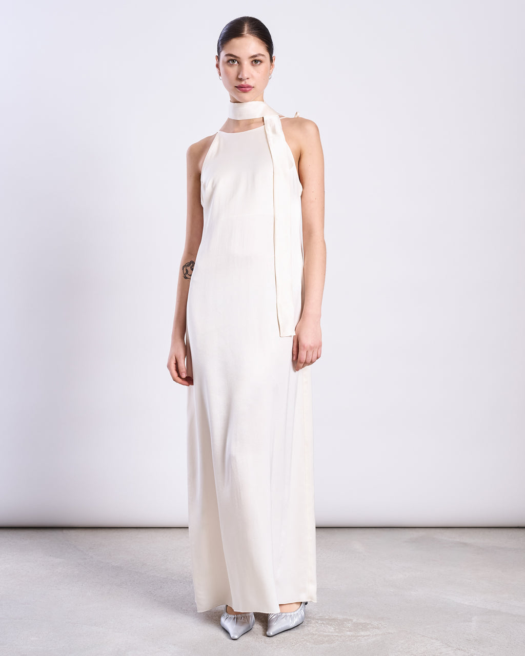 A woman stands against a plain background in a sleeveless, floor-length white vegan silk gown with a high neckline, styled with the jan-n-june SILKY SCARF PIURA OFFWHITE draped at her neck. Her hair is pulled back and she wears silver pointed-toe shoes.