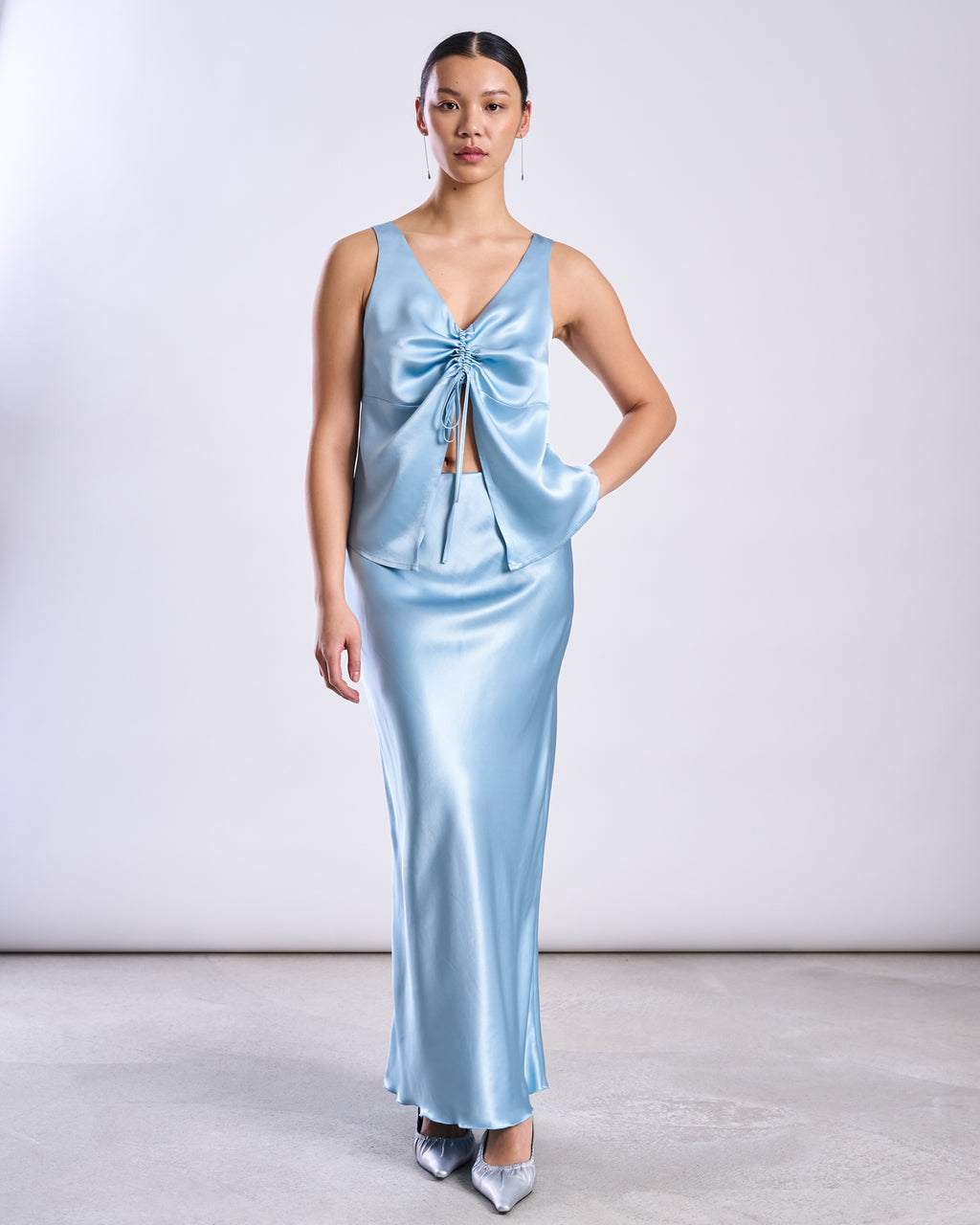 A woman poses against a plain background in a shiny, light blue jan-n-june MAXI SKIRT SERIN RETRO BLUE with a matching satin sleeveless top featuring a front tie. She wears pointed silver heels and has her hair pulled back.