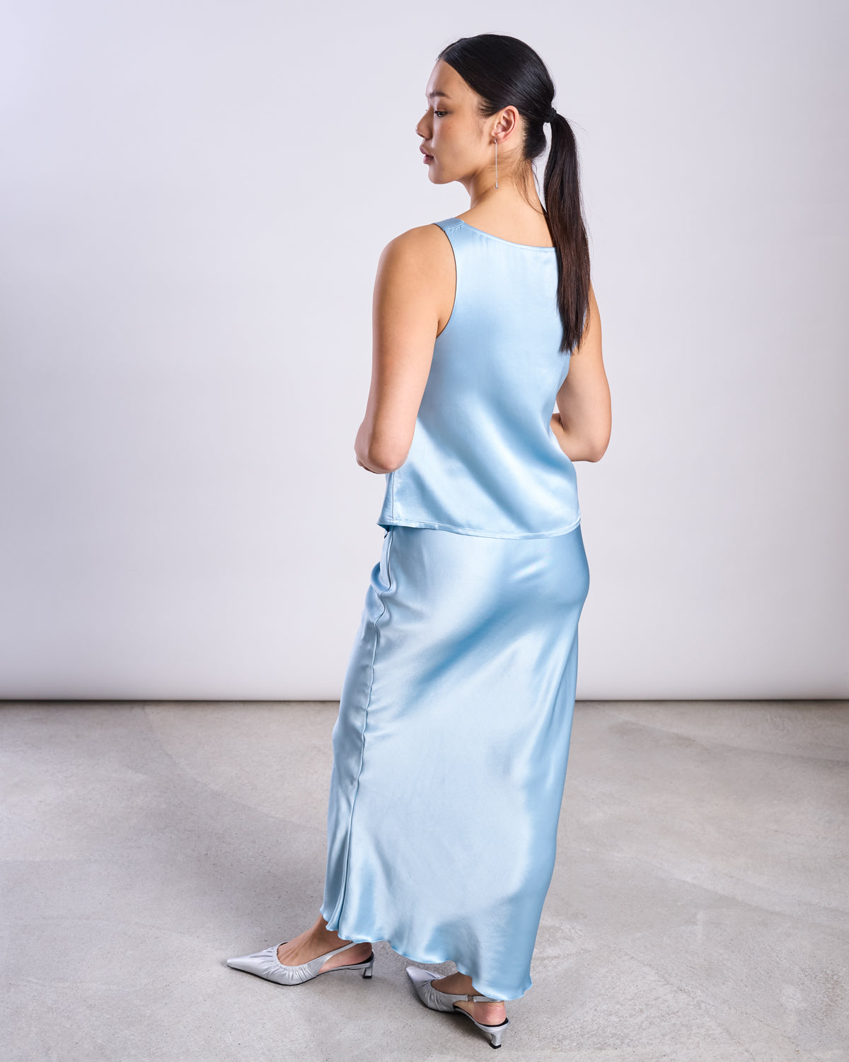 A woman in jan-n-june’s MAXI SKIRT SERIN RETRO BLUE stands on a gray floor, turned slightly away from the camera. She wears a matching sleeveless top, silver heels, and has her dark hair tied back—a modern look.