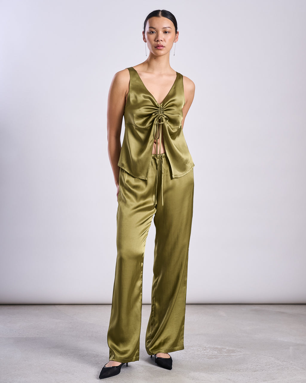 A person stands against a plain light background wearing a shiny woody olive RUFFLED V-NECK TOP TALARA by jan-n-june with matching wide-leg pants and black pointed-toe shoes.
