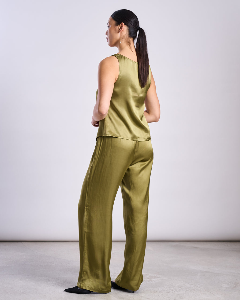 A woman with a low ponytail stands on a light gray floor against a plain background, wearing jan-n-junes LOOSE SILKY PANTS FRIS in woody olive along with a matching sleeveless vegan silk top.