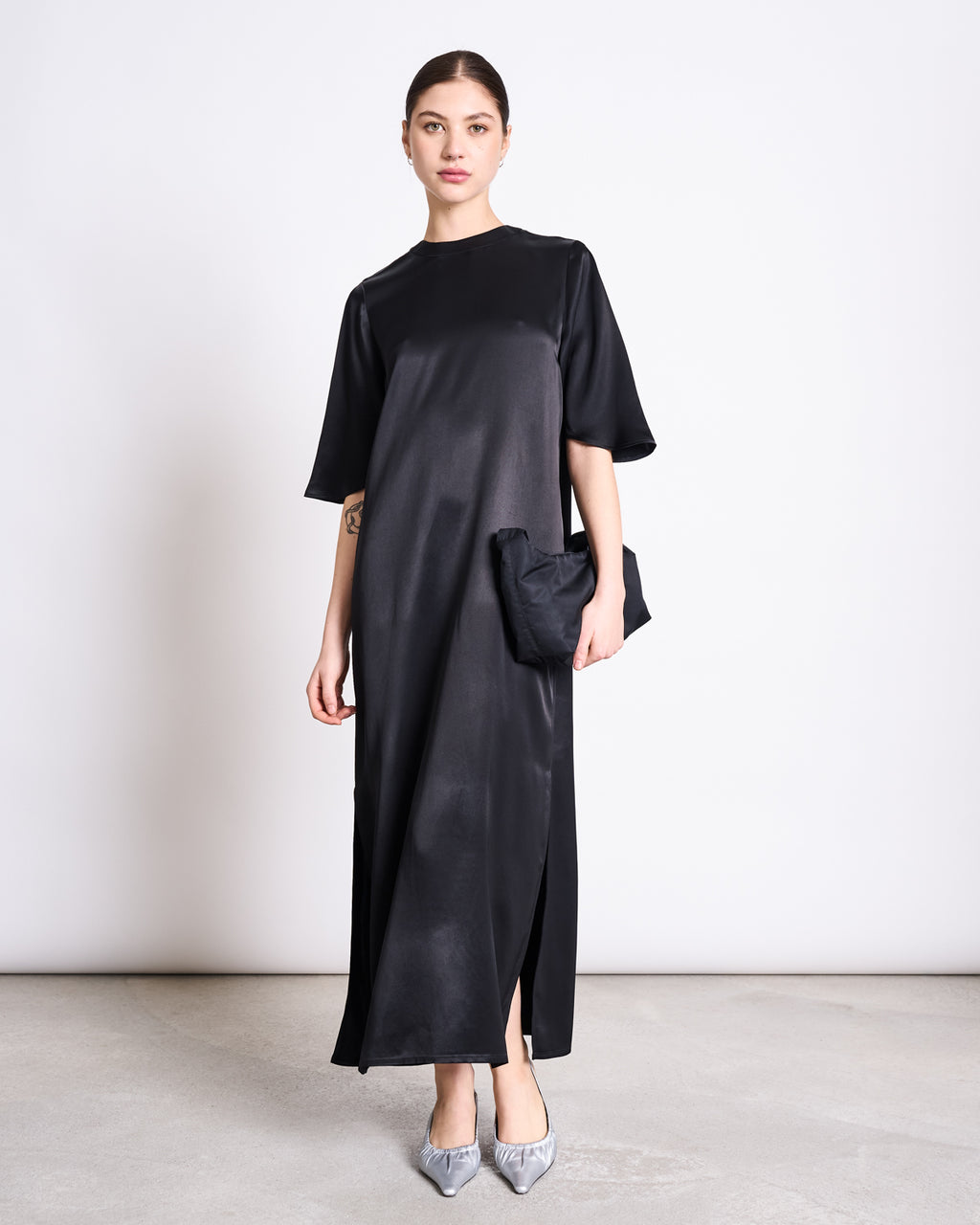 A woman stands against a plain wall wearing the jan-n-june SILKY T-SHIRT DRESS CALEU BLACK—a flowy, black satin dress with a round neck, short sleeves, and side slit. She pairs it with a black clutch and gray pointed-toe heels with bows.