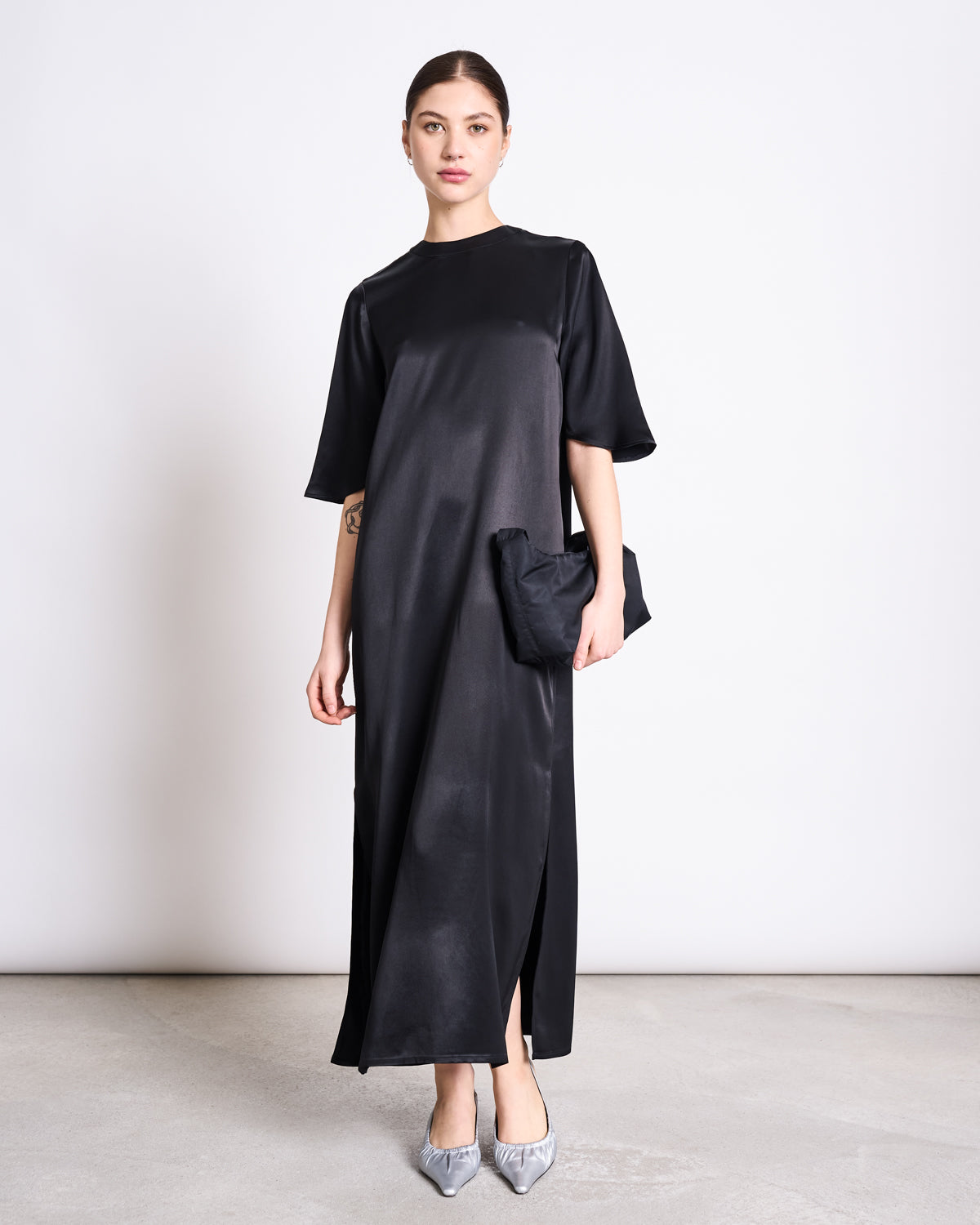 A woman stands against a plain wall wearing the jan-n-june SILKY T-SHIRT DRESS CALEU BLACK—a flowy, black satin dress with a round neck, short sleeves, and side slit. She pairs it with a black clutch and gray pointed-toe heels with bows.