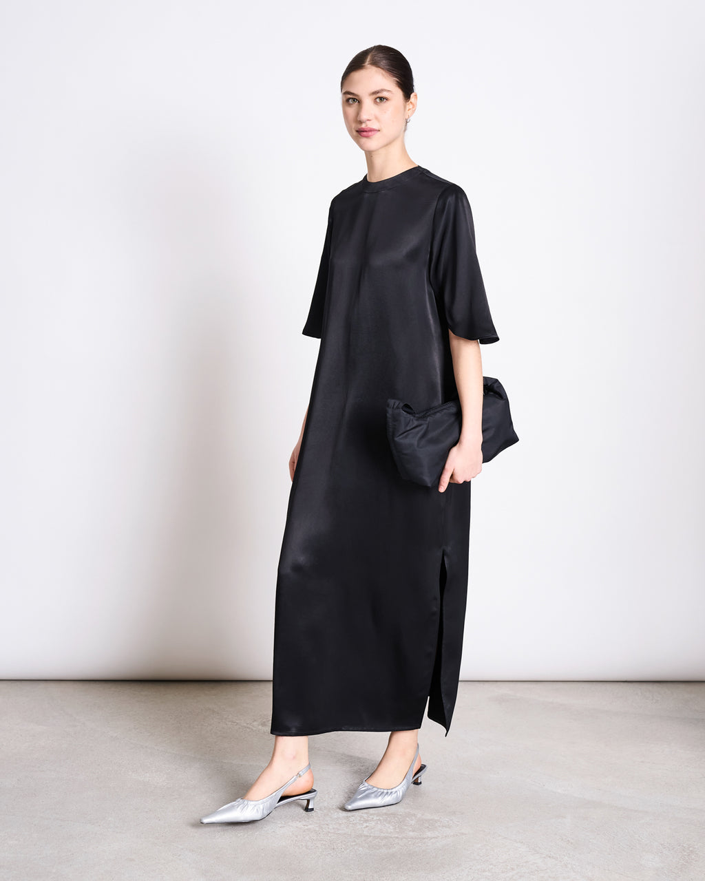 A woman stands against a plain background wearing the SILKY T-SHIRT DRESS CALEU BLACK by jan-n-june, a relaxed fit satin midi dress with short sleeves. She holds a black clutch, wears pointed silver heels, and has her hair neatly tied back.