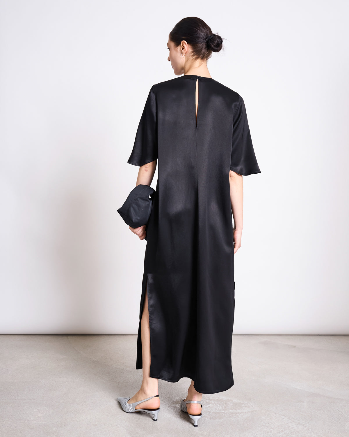 A woman stands facing away, wearing the SILKY T-SHIRT DRESS CALEU BLACK by jan-n-june—a relaxed fit, shin-length satin dress with short sleeves and a back slit. She holds a black clutch, wears low pointed heels, and her dark hair is in a bun.